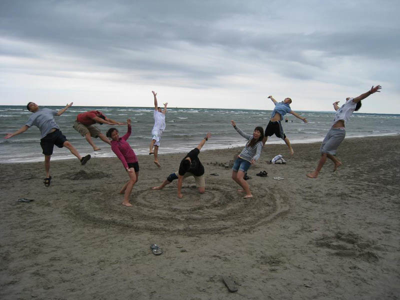 Imagem de pessoas na praia com homem ao centro fingindo dar soco na areia e todos pulando com o impacto.