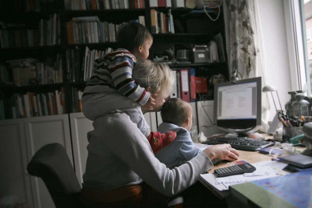 Foto mulher com dois filhos pendurados nela no Home Office.