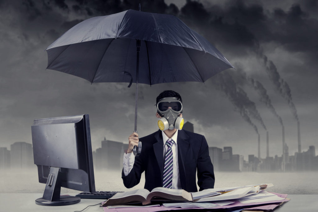 Homem de terno e gravata em frente a computador usando máscara de gás e guarda-chuvas.